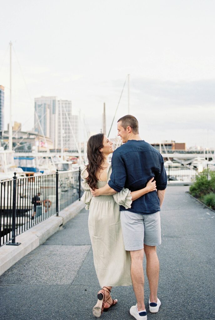 couple walk away from the camera with arms around each other's backs looking at one another in a harbor 