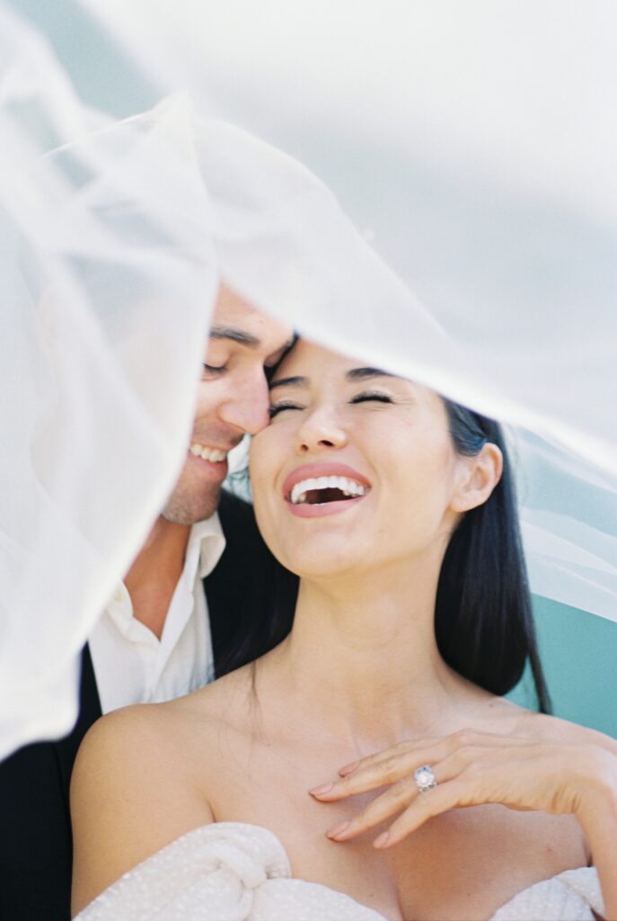 bride laughs as groom talks in her ear, a veil flys above them