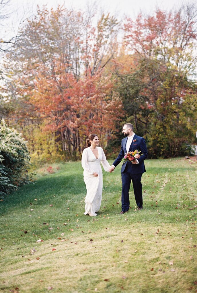 bride and groom walk together through field with iconic New England fall colors on the trees