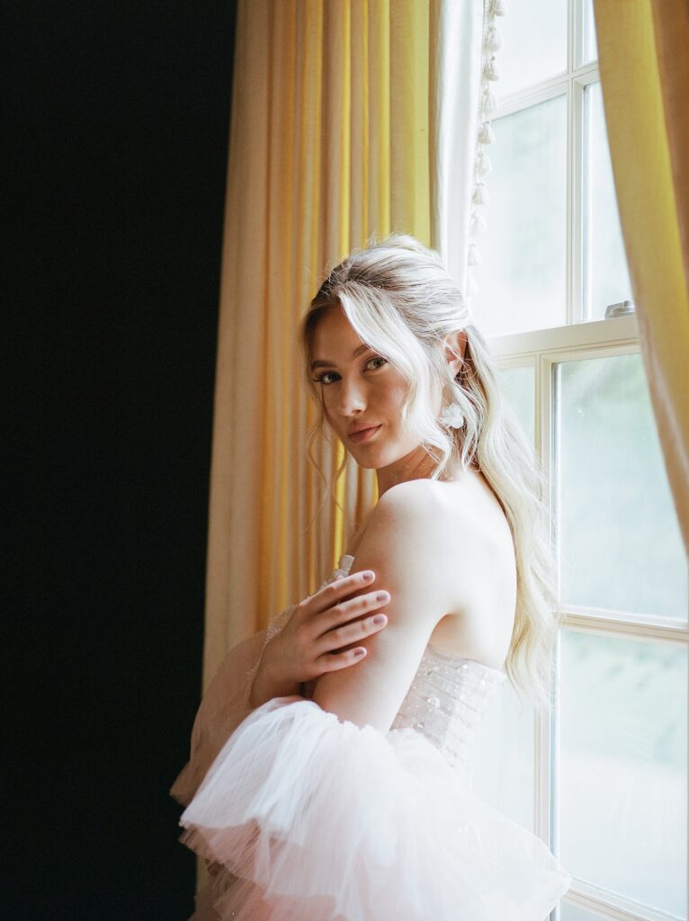 bride touches shoulder while looking half profile at the camera in front of a window with yellow curtains