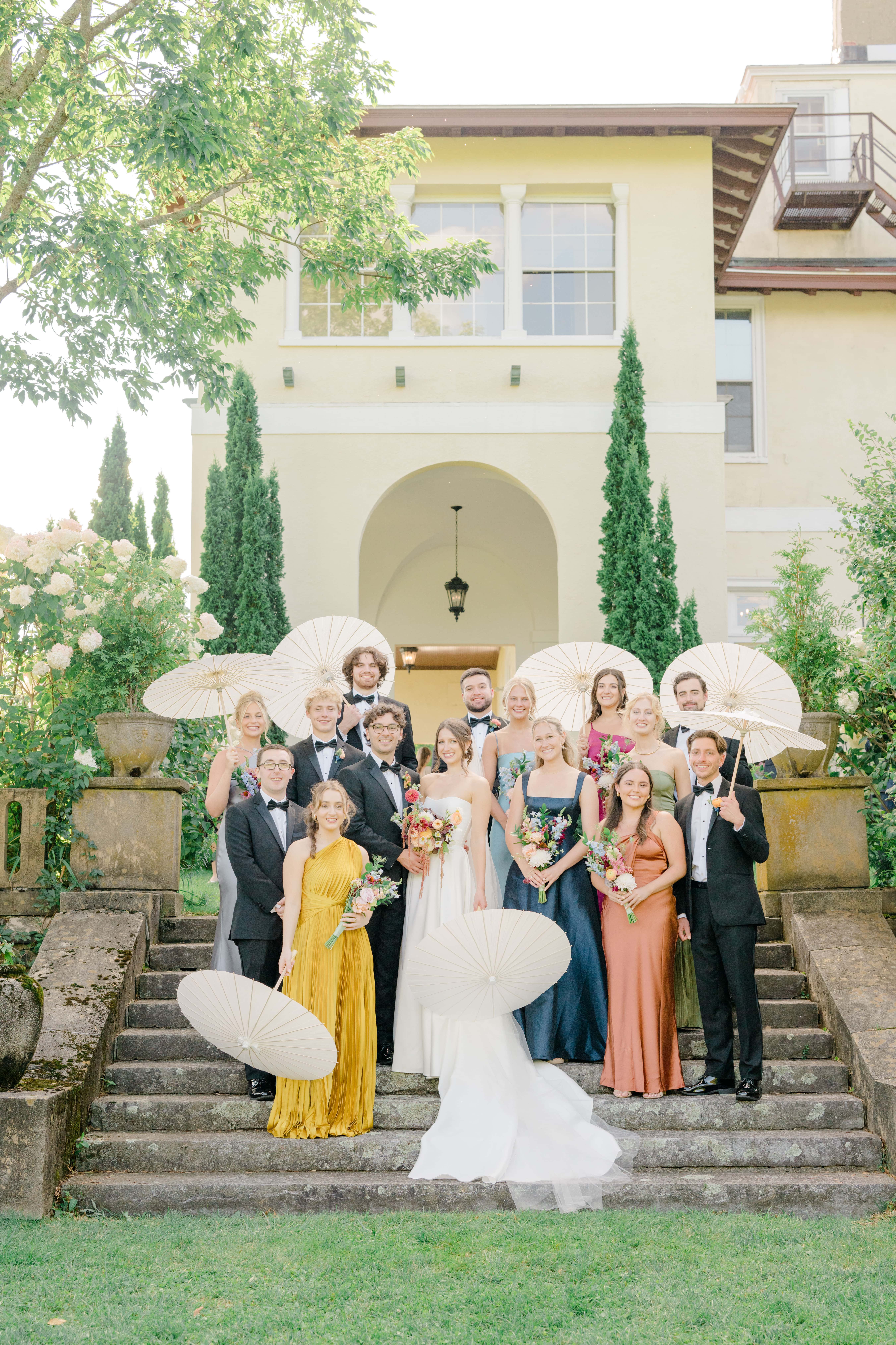 Bridal party gathered on the stone steps at Aldworth Manor, holding white parasols and wearing a mix of colorful dresses and tuxedos. The group is joyful, surrounded by greenery and flowers, with the venue's grand architecture in the background.