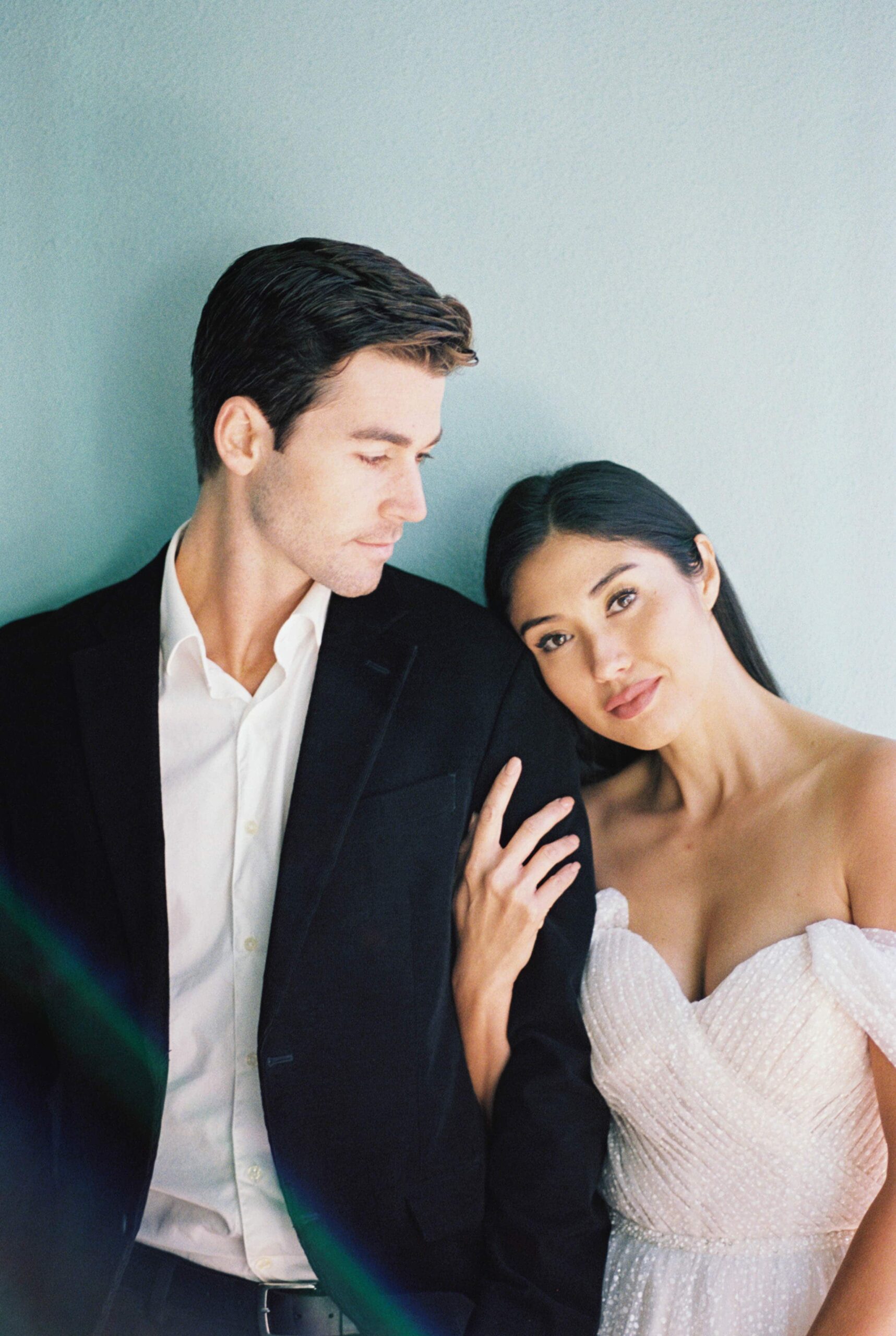 A romantic couple portrait in Charleston, SC, featuring the groom in a black suit and the bride in a beaded white wedding dress, standing closely together with soft smiles against a pastel blue wall.