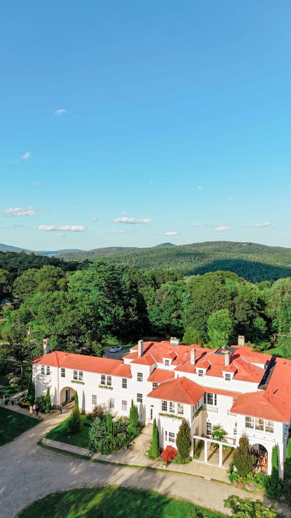 Aerial view of a historic white estate with red roofs surrounded by rolling green hills, showing the secluded setting of this Harrisville, New Hampshire wedding venue.