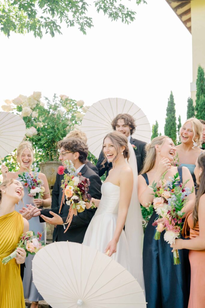 Bride and groom laugh with their wedding party beneath white parasols and colorful flowers, celebrating outdoors at Aldworth Manor in Harrisville, NH.