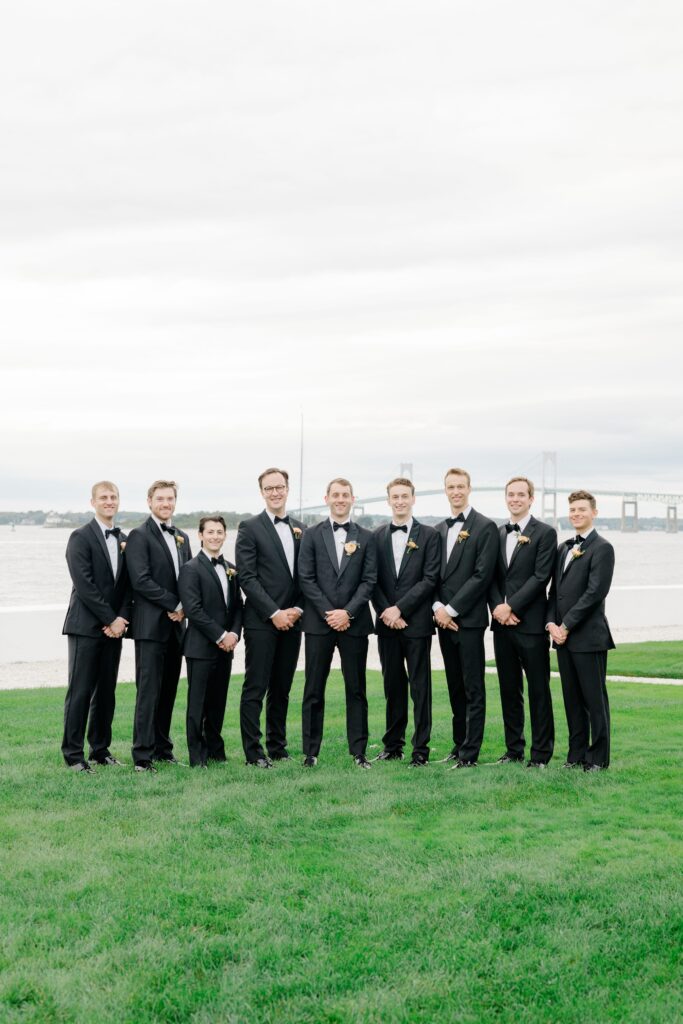 Groomsmen in classic black tuxedos posing on Belle Mer lawn overlooking the water.