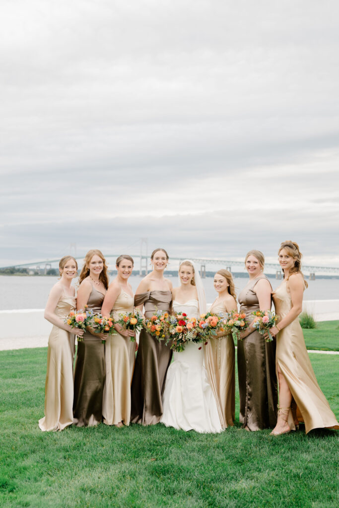 Bridal party of bridesmaids in neutral dresses with colorful bouquets on Belle Mer lawn overlooking Newport Harbor.