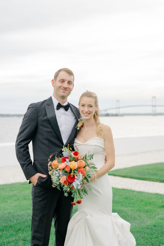 Bride and groom portrait on Belle Mer lawn with colorful bridal bouquet and coastal backdrop.
