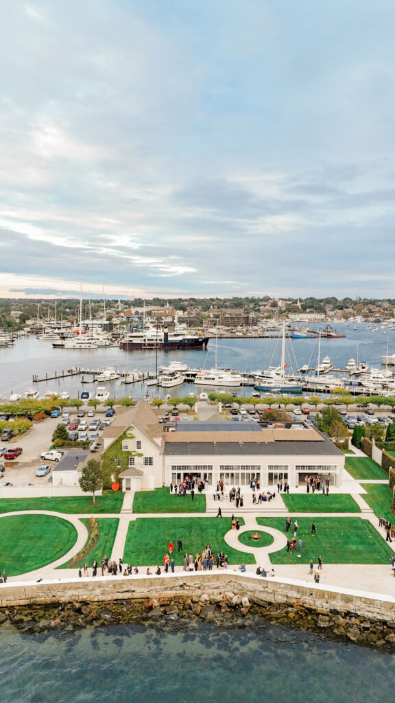 Aerial view of Belle Mer venue and Newport Harbor with wedding guests gathered on lawn for cocktail hour.