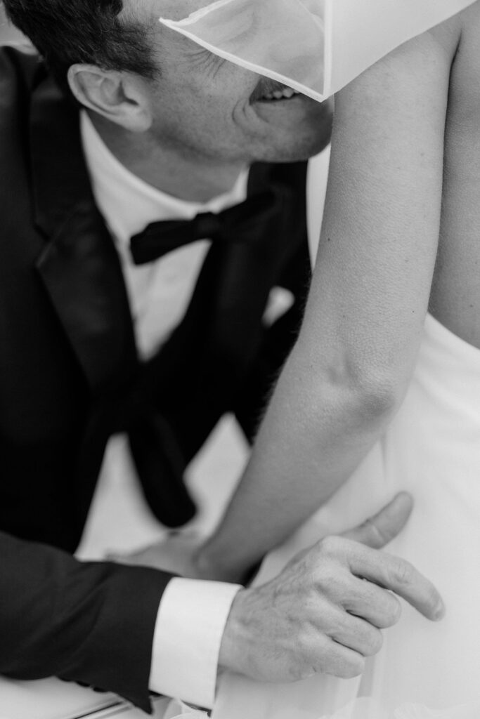 Groom kissing bride's shoulder beside white vintage Mercedes — stunning open-back halter dress with ruffled tulle skirt and bow from Gown Boutique of Charleston