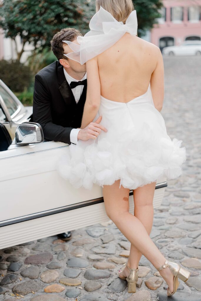 Bride in open-back halter tulle mini dress with bow detail beside vintage white Mercedes on Rainbow Row cobblestone streets in Charleston SC