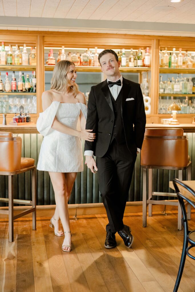 Stylish couple posing at the bar inside a stunning Charleston SC hotel during a downtown couples photography session — bride in white mini dress, groom in black tuxedo