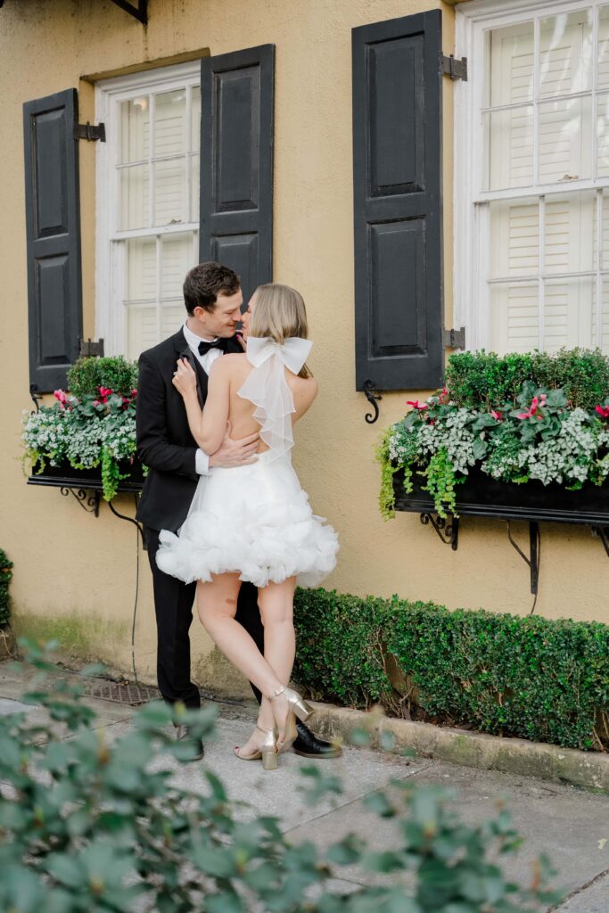 Close-up detail of bride in white ruffled mini dress and gold strappy heels beside groom in black tuxedo against a warm yellow stucco wall in downtown Charleston SC