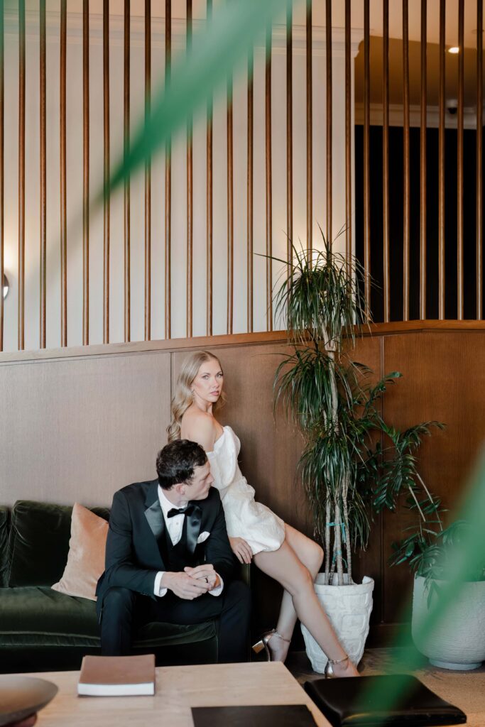 Editorial couples portrait inside Hotel Emeline in downtown Charleston SC — bride in white off-shoulder mini dress perched behind groom on emerald velvet sofa