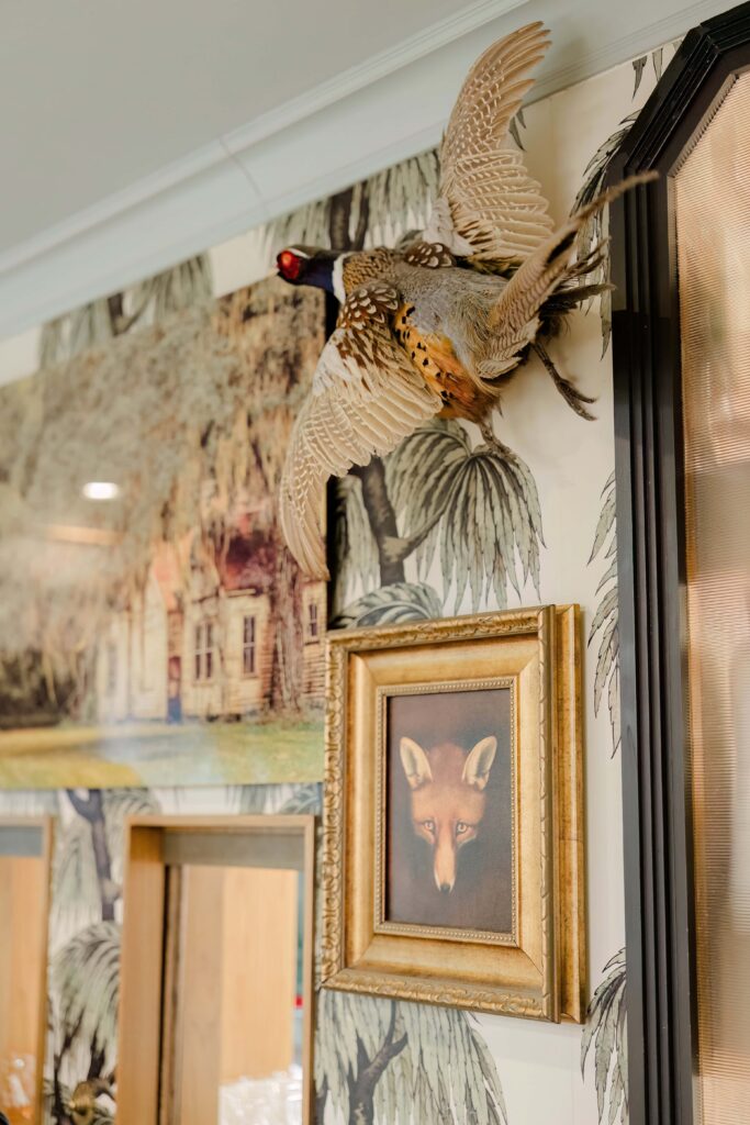 Stunning eclectic interior decor at Hotel Emeline Charleston SC — mounted pheasant taxidermy above gold-framed fox portrait against botanical wallpaper