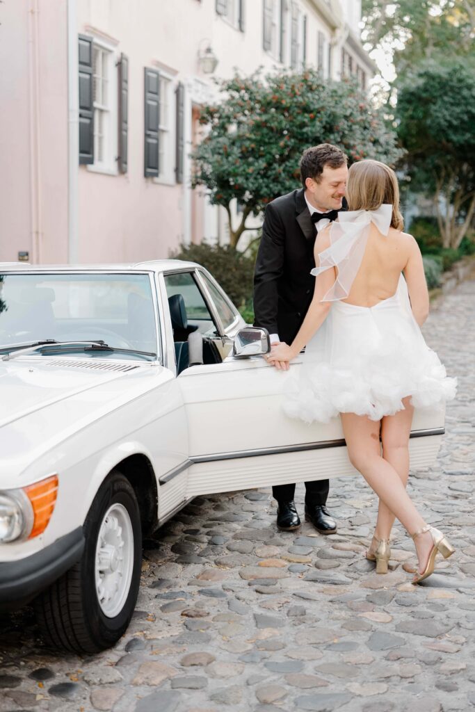 Joyful couple laughing together against a white vintage Mercedes on cobblestone streets in downtown Charleston SC — ivy-covered stone arch and red door behind them