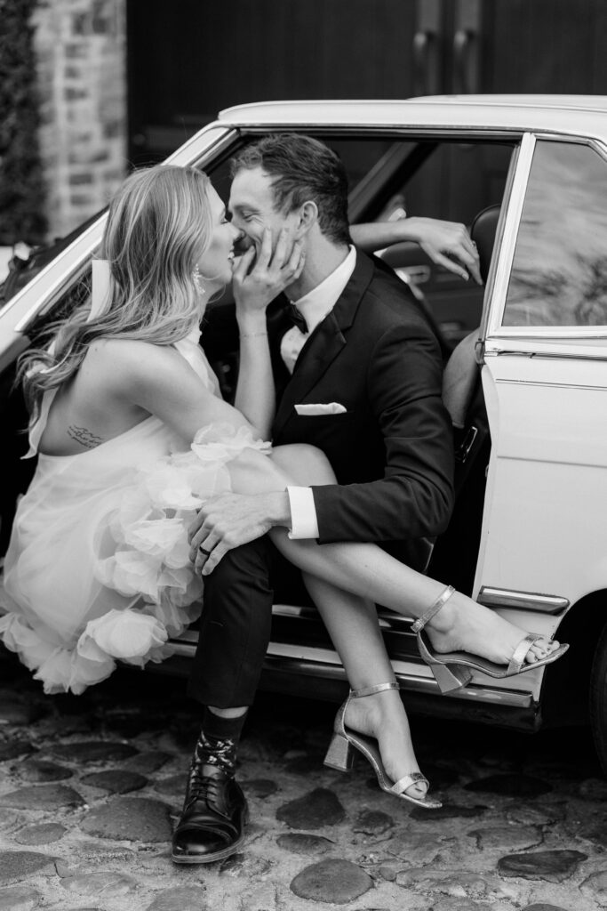 Black and white photo of couple kissing beside a white vintage Mercedes on cobblestone streets in downtown Charleston SC — romantic couples photography