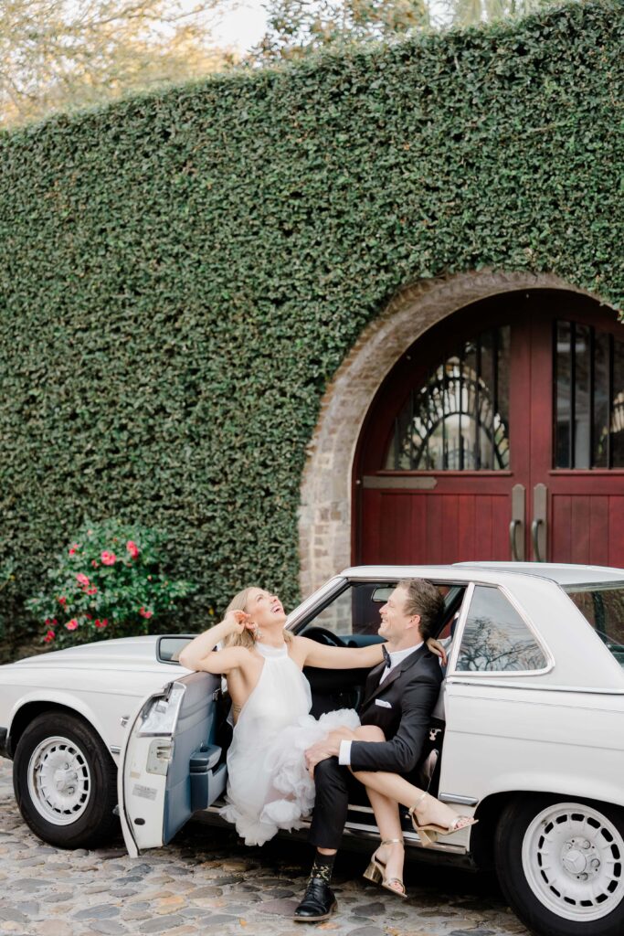 Laughing couple sitting in the open door of a white vintage Mercedes convertible during a Charleston SC couples session — ivy wall and red arched door backdrop