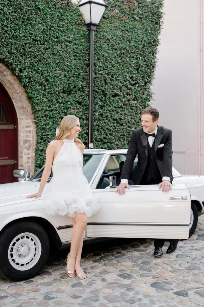 Bride and groom smiling at each other beside a white vintage Mercedes on historic cobblestone streets in downtown Charleston SC