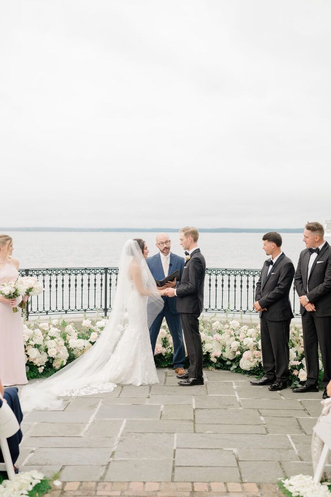 The couple exchange vows on a stone terrace overlooking the water, surrounded by white florals and loved ones at one of the most scenic oceanfront weddings in New England.