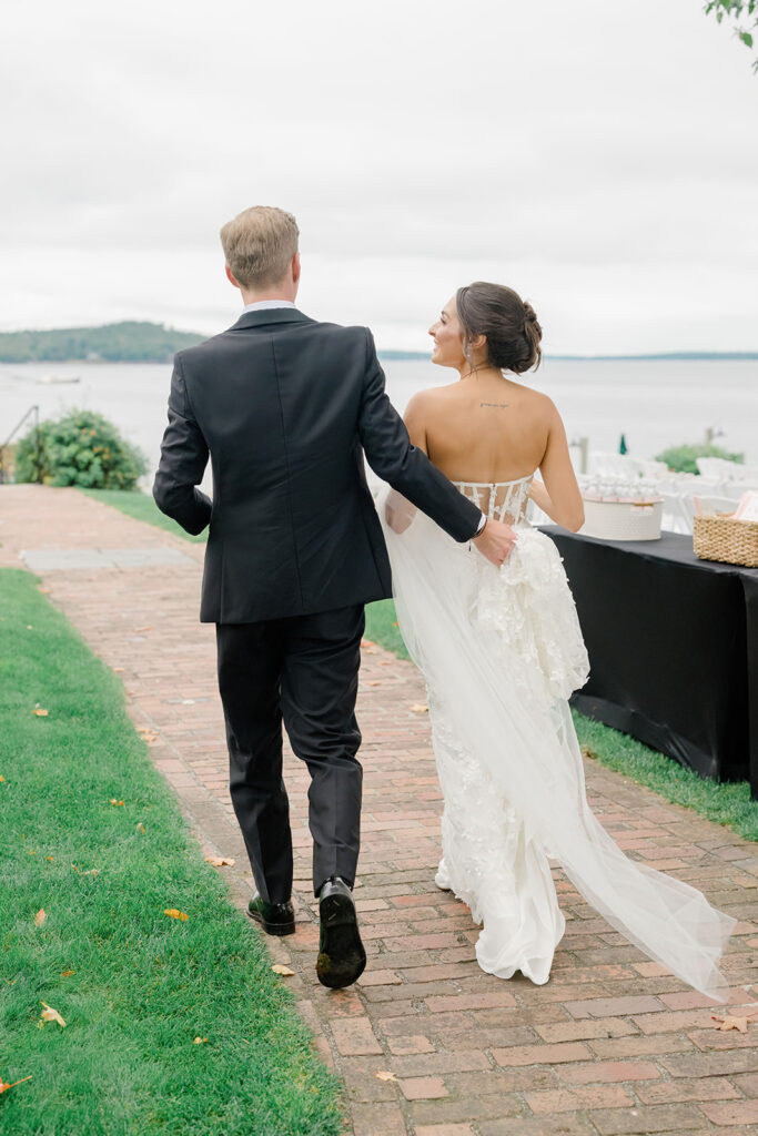 Newlyweds walk hand in hand along a brick path beside the water, blending quiet portraiture with the scenery of a waterfront New England wedding.