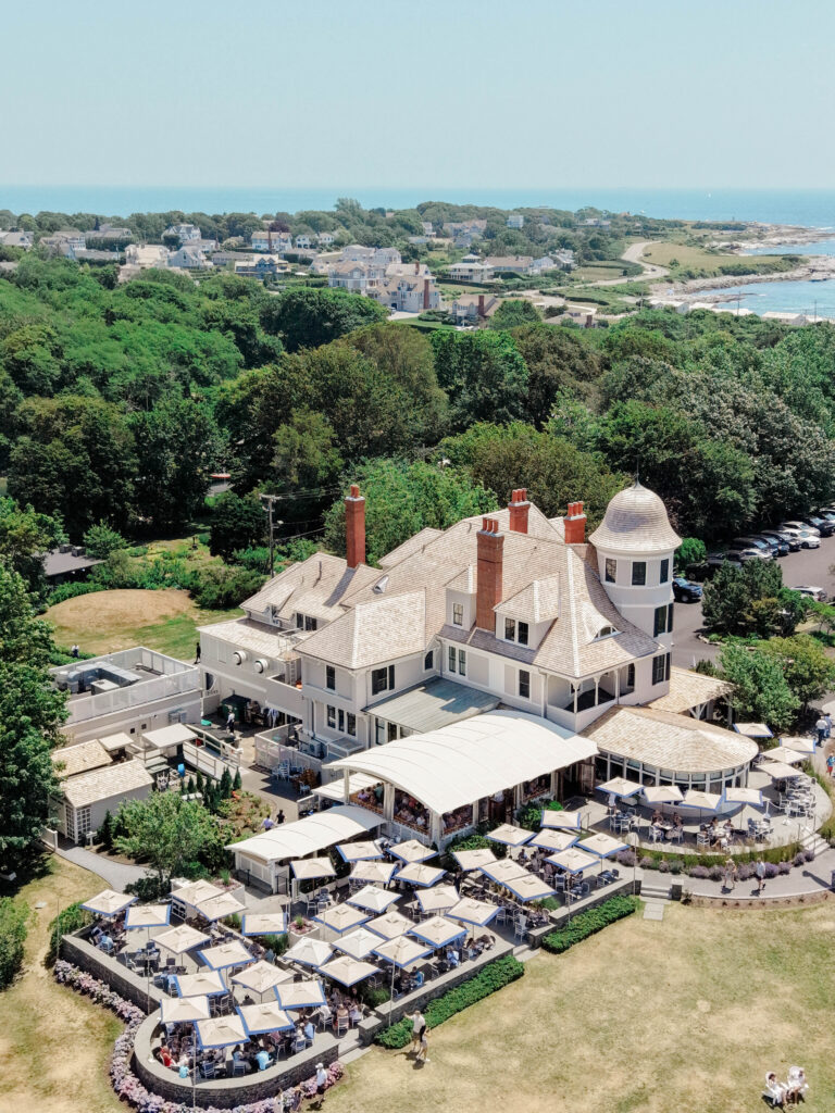 Aerial view of a grand coastal estate with outdoor dining terraces, surrounding greenery, and ocean views, showcasing Newport Rhode Island wedding venues.