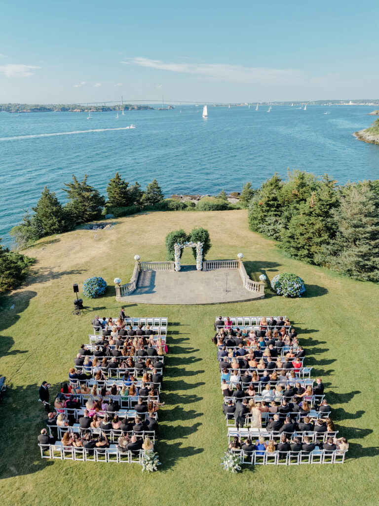 Aerial view of an outdoor wedding ceremony arranged on a seaside lawn, with blue water stretching beyond the guests at a coastal wedding venue in New England.
