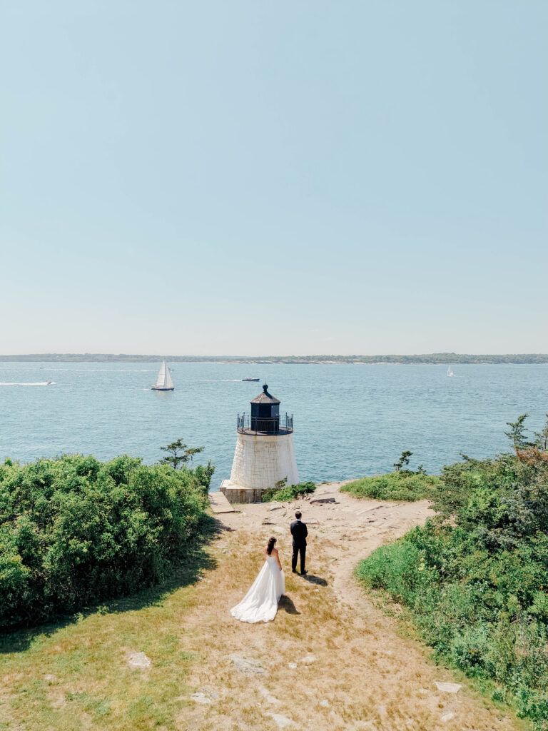 A wide portrait of the couple walking toward the lighthouse highlights the dramatic coastline and expansive waterfront views. One of the most iconic photo locations for Castle Hill Inn wedding photos.