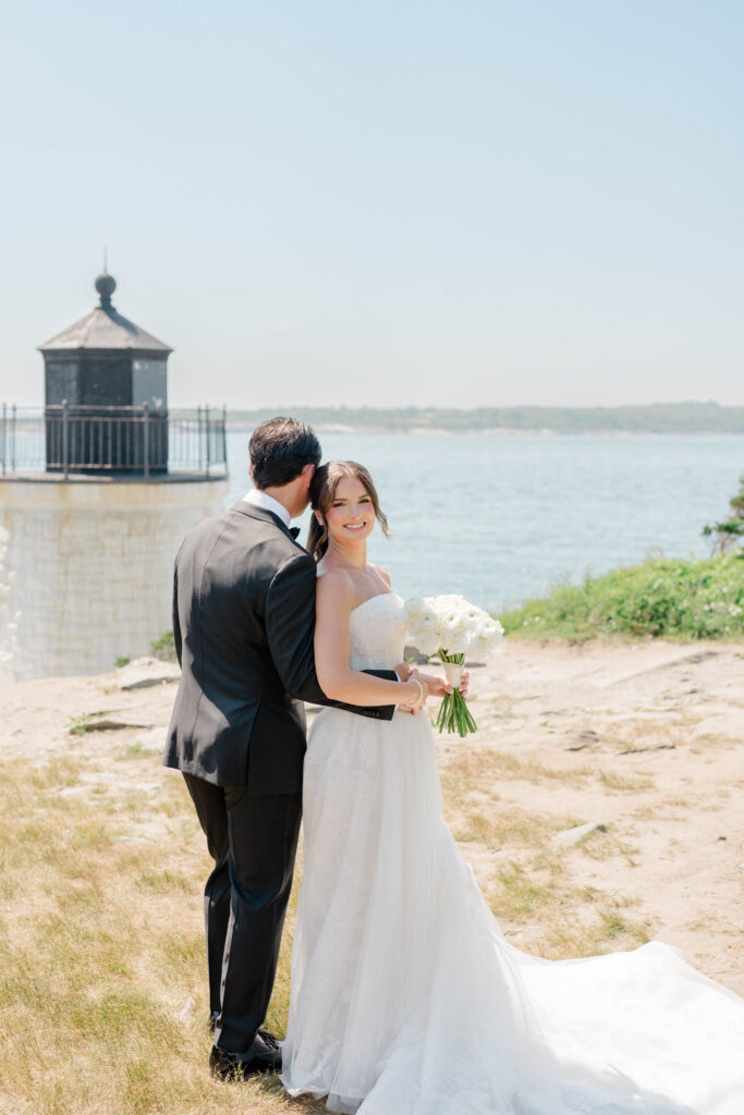 The newlyweds embrace beside the iconic lighthouse with sweeping coastal views stretching behind them. A timeless portrait from a Castle Hill Inn wedding on the Newport shoreline.