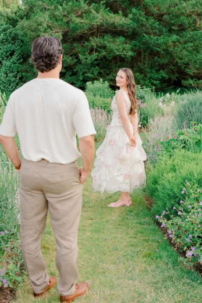 The couple walks hand in hand through a flower-lined garden path, creating a sense of movement and anticipation. A romantic perspective on Connecticut engagement photo locations.
