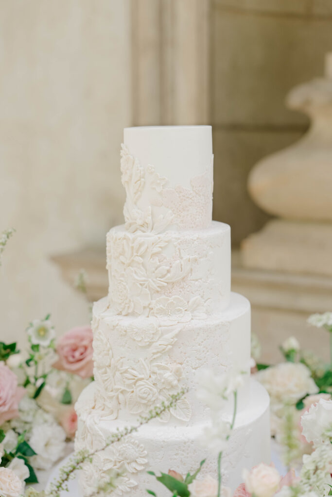Four-tier white wedding cake features intricate floral detailing surrounded by blush blooms, an elegant detail suited to a Vanderbilt Museum wedding reception.