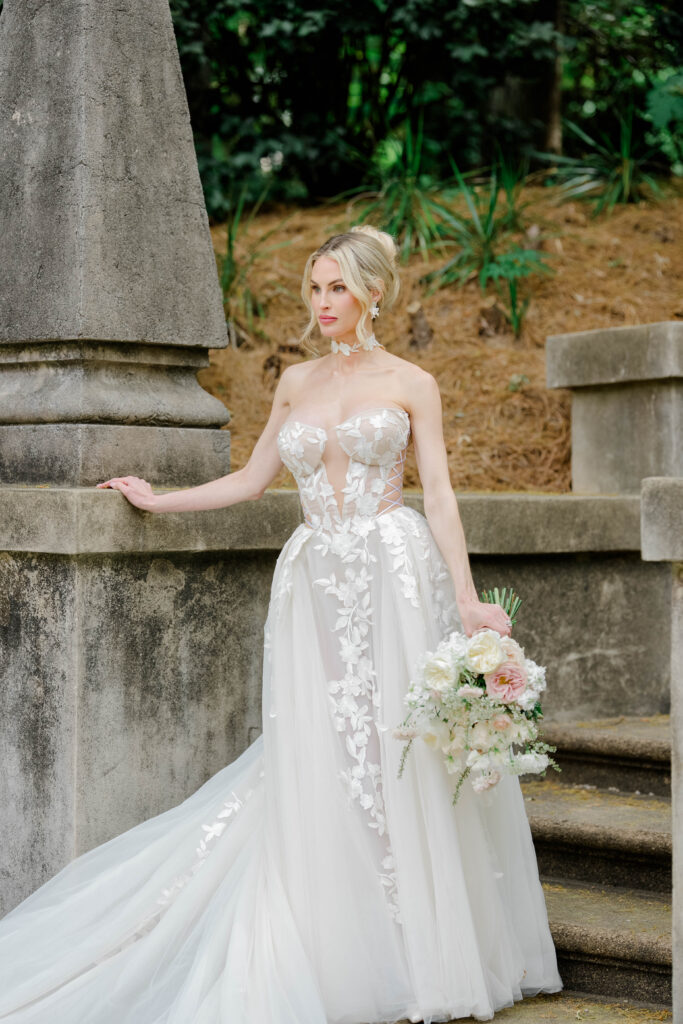 The bride stands gracefully on the historic stone staircase holding a pastel bouquet, with the estate providing a refined backdrop. Fashion-forward bridal portraits inspired by a Swan House wedding.