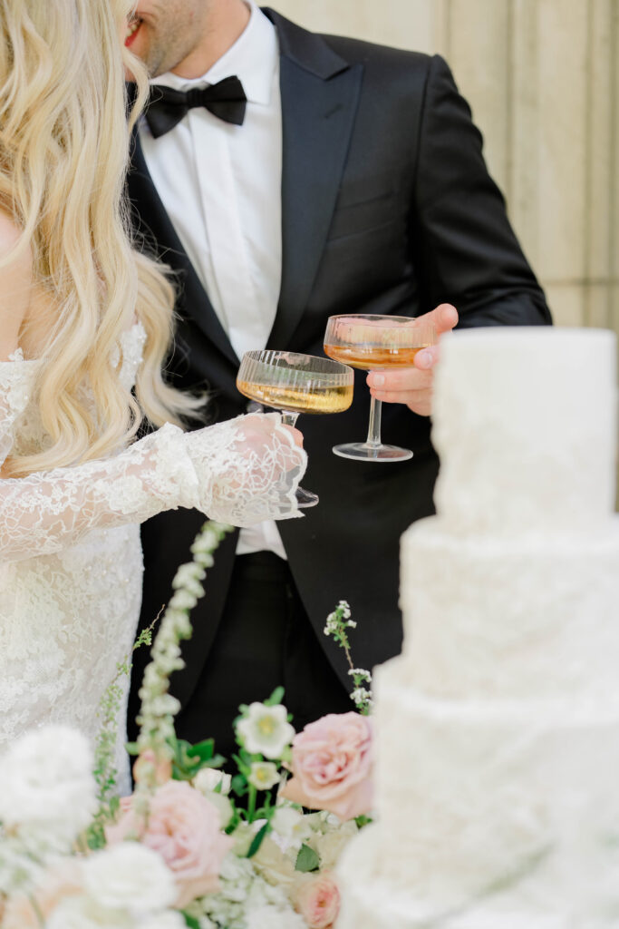The couple shares a champagne toast beside their wedding cake, surrounded by delicate florals and reception details. Couture celebration inspiration from a luxury Atlanta wedding editorial.