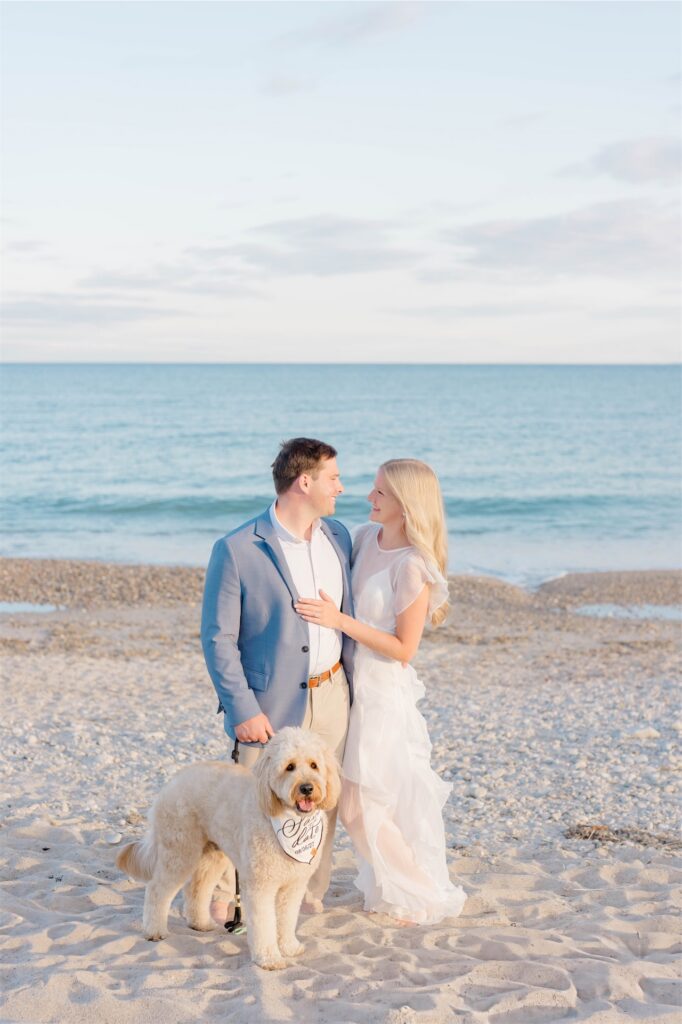 Molly and Anthony posing with their dog on a Rhode Island beach, with calm ocean water in the background.