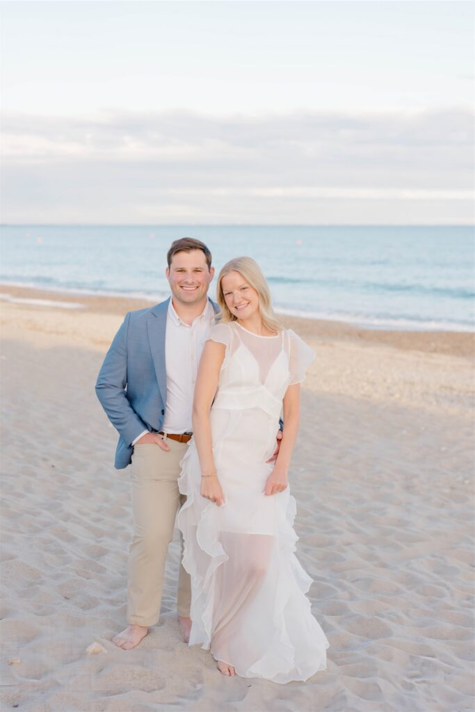 Molly and Anthony standing barefoot together on the sandy shoreline with the ocean behind them.