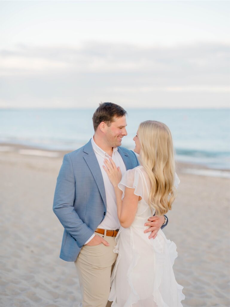 Molly and Anthony smiling at each other beside the ocean during their Rhode Island beach engagement photos.