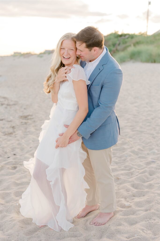 Anthony embracing Molly from behind as she laughs during their beach engagement session in Wakefield, Rhode Island.