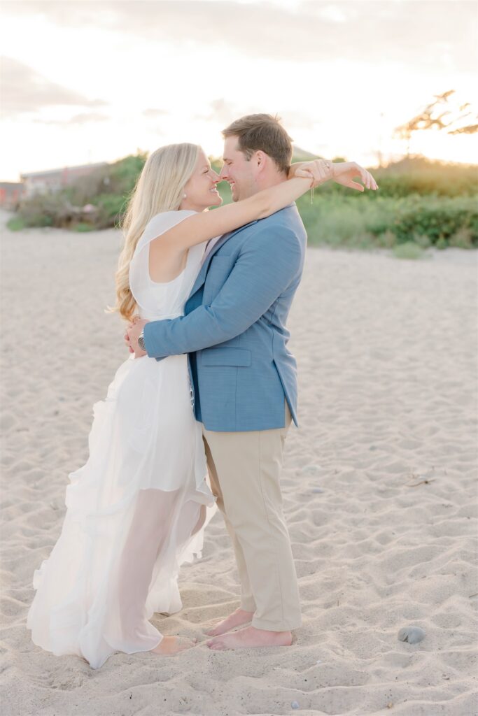 Molly and Anthony embracing barefoot on the sand with coastal grasses and warm evening light behind them.