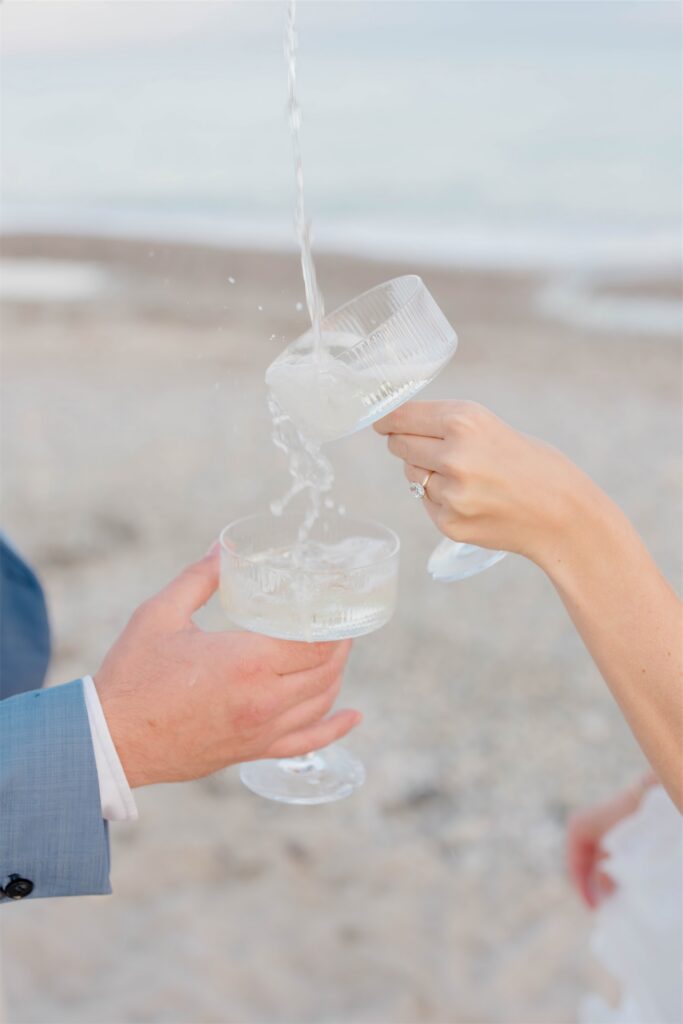 Close-up of Molly and Anthony pouring champagne into coupe glasses during their beach engagement session.