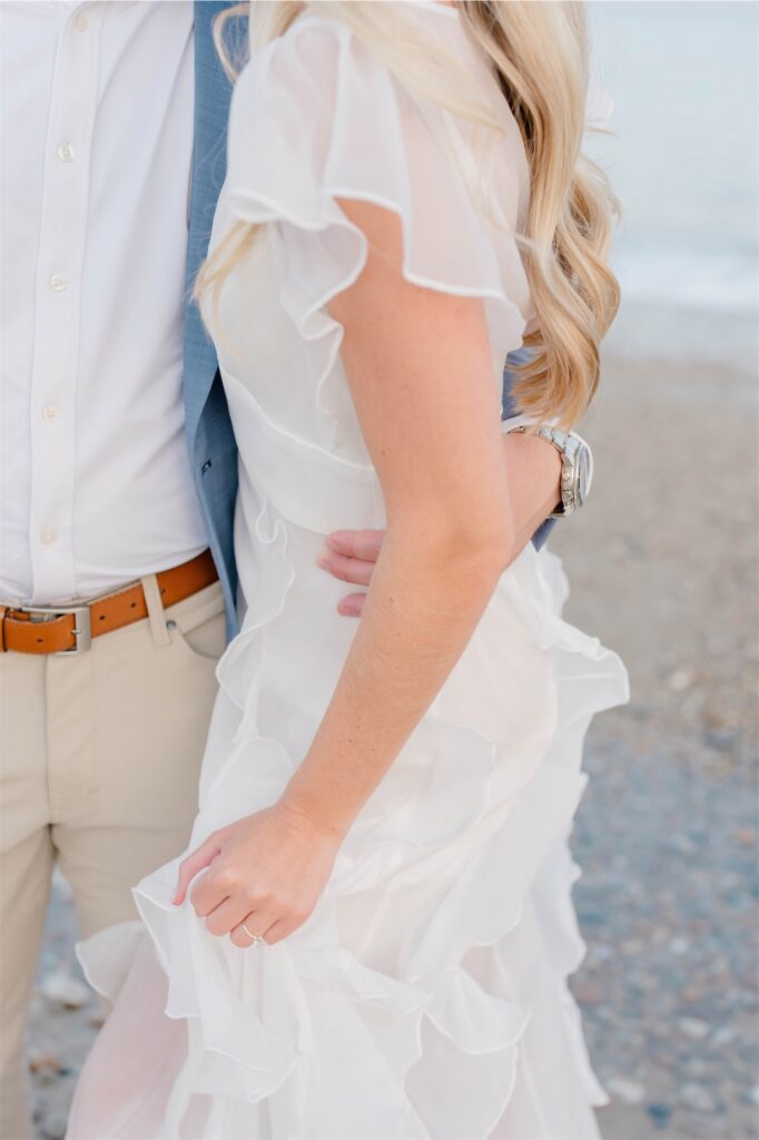 Close-up of Molly's white ruffled dress and engagement ring as Anthony holds her waist on the beach.