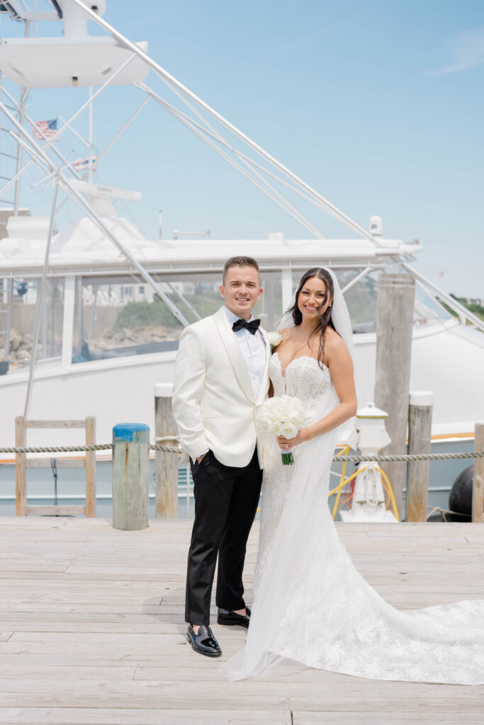 Bride and groom posing on the marina dock with a fishing boat behind them at Wychmere Harbor.